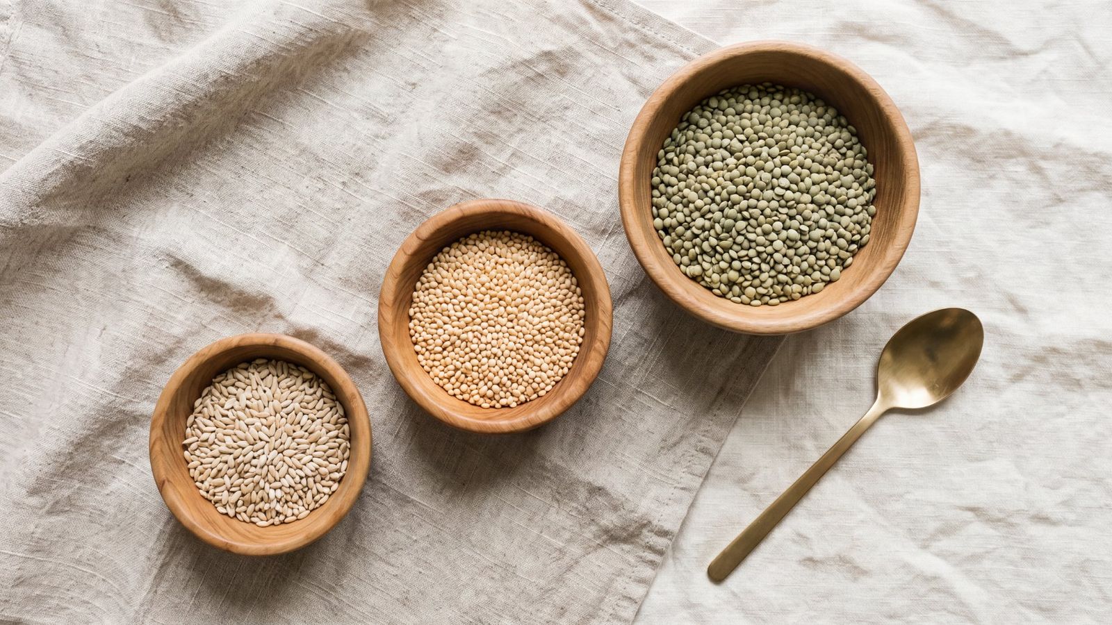 Three wooden bowls of grains of different sizes on linen, arranged in order
